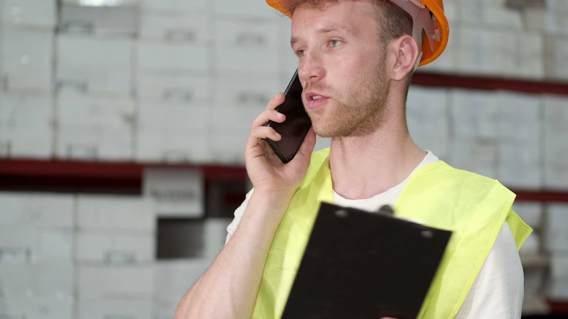 A site manager in a hard hat on the phone, holding a clipboard