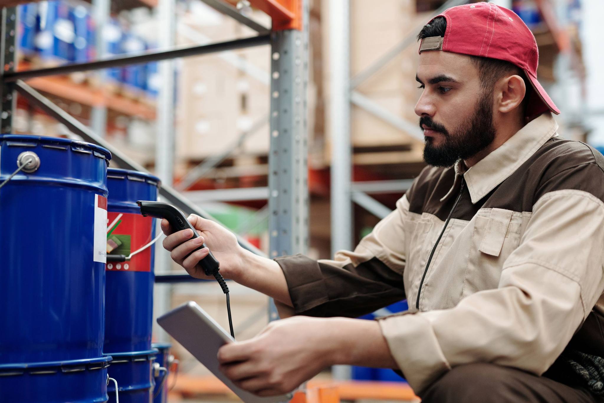 A man in work overalls scanning the label on a drum with a handheld scanner, holding a tablet
