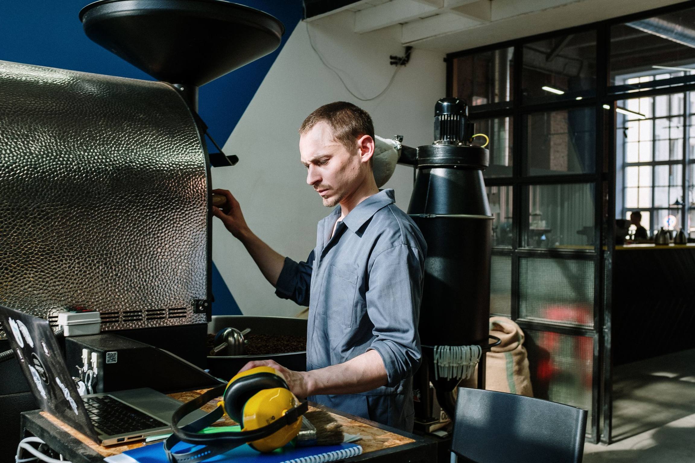 A man adjusting a machine by hand, with a laptop and ear defenders on the bench beside him