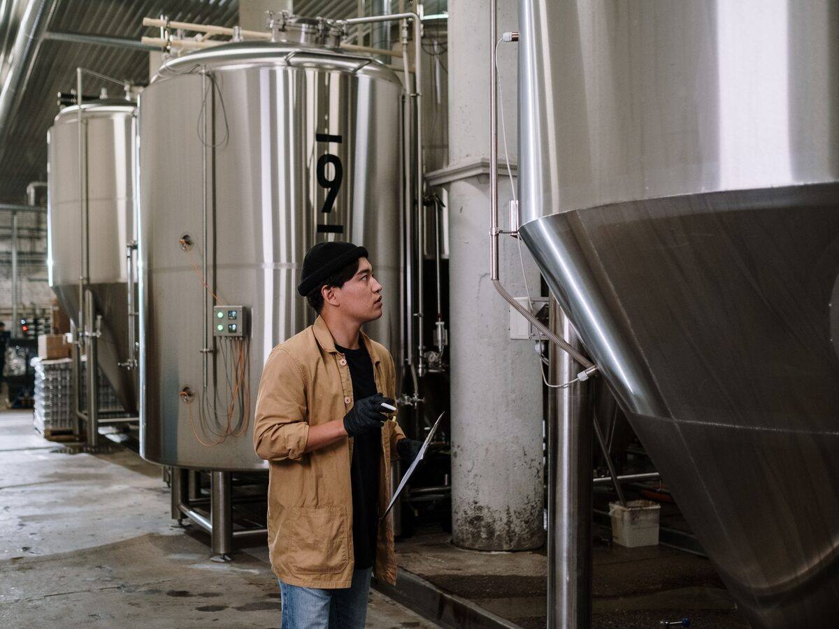 A man with a clipboard and pen looking up at tall stainless steel tanks in a production plant